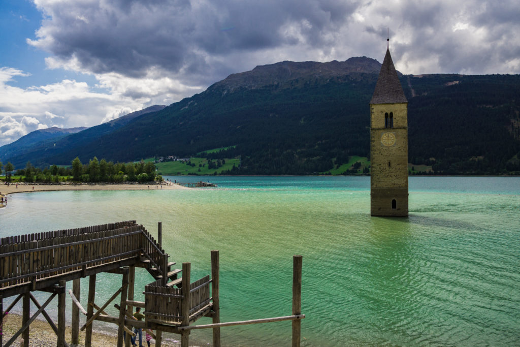 Das Foto zeigt den Kirchturm im Reschensee - eines der Wahrzeichen des Vinschgaus - und einen Holzsteg als Fotopunkt.