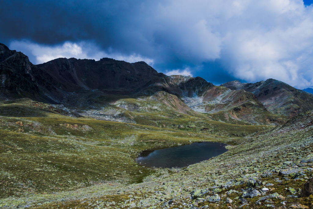 Das Bild zeigt einen kleinen, klaren Bergsee im unwirtlichen Gelände unweit des Niederjöchls mit dramatischen Wolken.